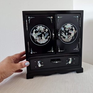 May include: A black wooden jewelry box with a hinged lid and a drawer. The lid features two circular inlays of mother-of-pearl with a floral design and birds. The drawer has a similar inlay with a floral design.