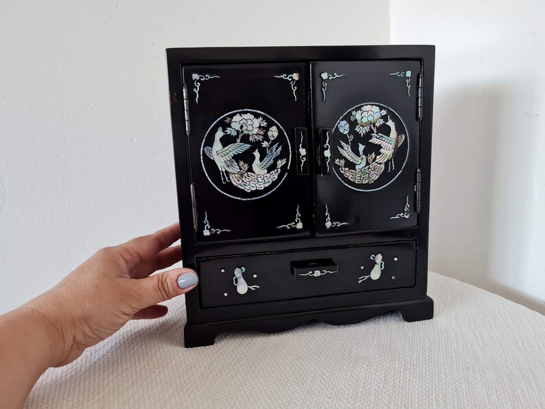 May include: A black wooden jewelry box with a hinged lid and a drawer. The lid features two circular inlays of mother-of-pearl with a floral design and birds. The drawer has a similar inlay with a floral design.