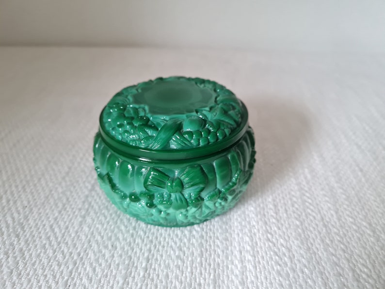 May include: A green glass trinket box with a carved design of flowers and bows. The lid is closed.