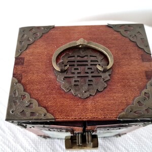 May include: A wooden box with a brass handle and decorative corners. The handle is shaped like a ring and has a Chinese character in the center. The box is closed.