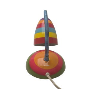 May include: A colorful desk lamp with a striped lampshade in red, yellow, blue, and green. The base is round with concentric circles of green, pink, orange, and yellow. A flexible blue neck connects the shade to the base.