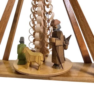 May include: A wooden Christmas pyramid with a carved shepherd figure, a sheep, and a tree made of curled wood. The pyramid is made of light brown wood and has a white base.