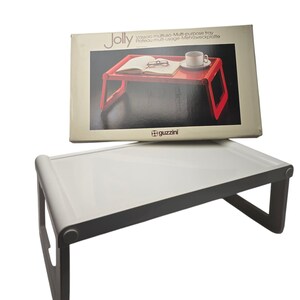 May include: A white and grey multi-purpose tray with a rectangular surface and angled legs. The tray is shown with its original box, which features a red tray with a book, glasses, and a cup.