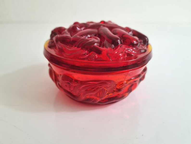 May include: A red glass trinket box with a lid featuring a raised design of intertwined figures. The box is round and has a smooth, polished finish.