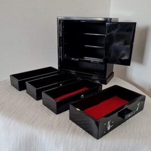 May include: Black wooden jewelry box with a hinged lid and four pull-out drawers. The drawers have red felt lining and are decorated with a floral design.