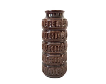 May include: A tall, brown ceramic vase with a geometric pattern. The vase has a cylindrical shape and is decorated with a repeating pattern of squares and rectangles.