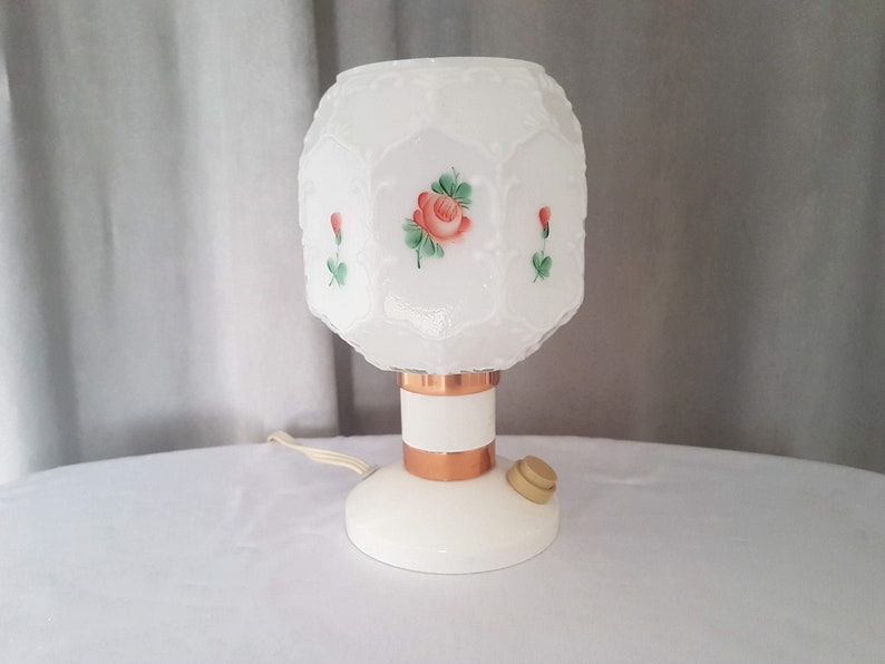May include: White table lamp with a frosted glass shade featuring a painted rose design. The shade has a geometric pattern and the base is copper colored.