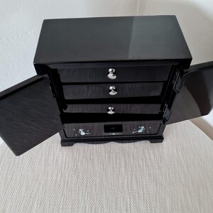 May include: Black wooden jewelry box with four drawers and two doors. The doors are open and the drawers are closed. The box has a decorative design on the front.