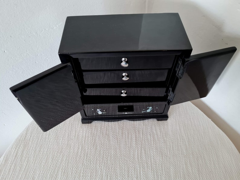 May include: Black wooden jewelry box with four drawers and two doors. The doors are open and the drawers are closed. The box has a decorative design on the front.