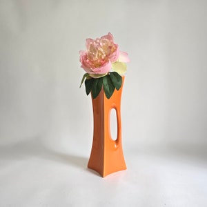 May include: A bright orange ceramic vase with a unique cutout design. The vase is filled with a single pink and white flower.