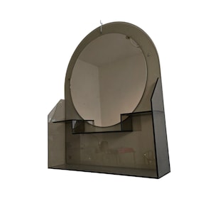 Op de afbeelding: Een vintage, gerookte glazen spiegel met een ronde spiegel en twee planken. De spiegel is aan de muur bevestigd met een haak.