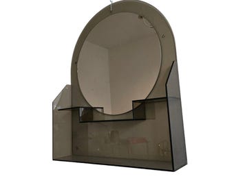 May include: A vintage, smoked glass vanity mirror with a round mirror and two shelves. The mirror is attached to a wall with a hook.