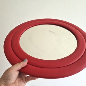 May include: A red round mirror with a double rim. The mirror is held in a hand.