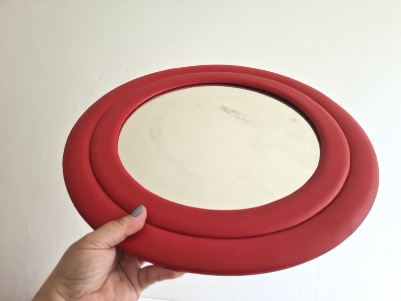 May include: A red round mirror with a double rim. The mirror is held in a hand.
