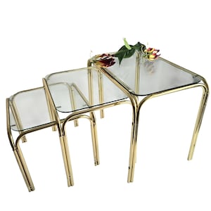 Pode incluir: Conjunto de três mesas de encaixe com tampos de vidro transparente e estruturas de metal dourado. As mesas estão dispostas em configuração escalonada, com um arranjo floral na mesa maior. As mesas têm uma estética retro e vintage.