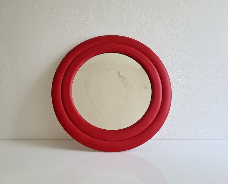 May include: A round red mirror with a double frame. The mirror has a simple, modern design.