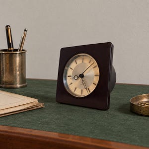 May include: A square desk clock with a dark brown frame and a circular metallic face. The clock features thin black hands and simple hour markers. The design is clean and functional, showcasing a polished finish on the frame and a reflective dial.