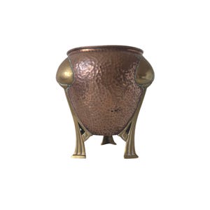 May include: A copper planter with a hammered texture, brass details, and three decorative legs. The planter has a rounded shape with a brass rim. The brass accents are on the sides and legs. The planter is suitable for indoor use.