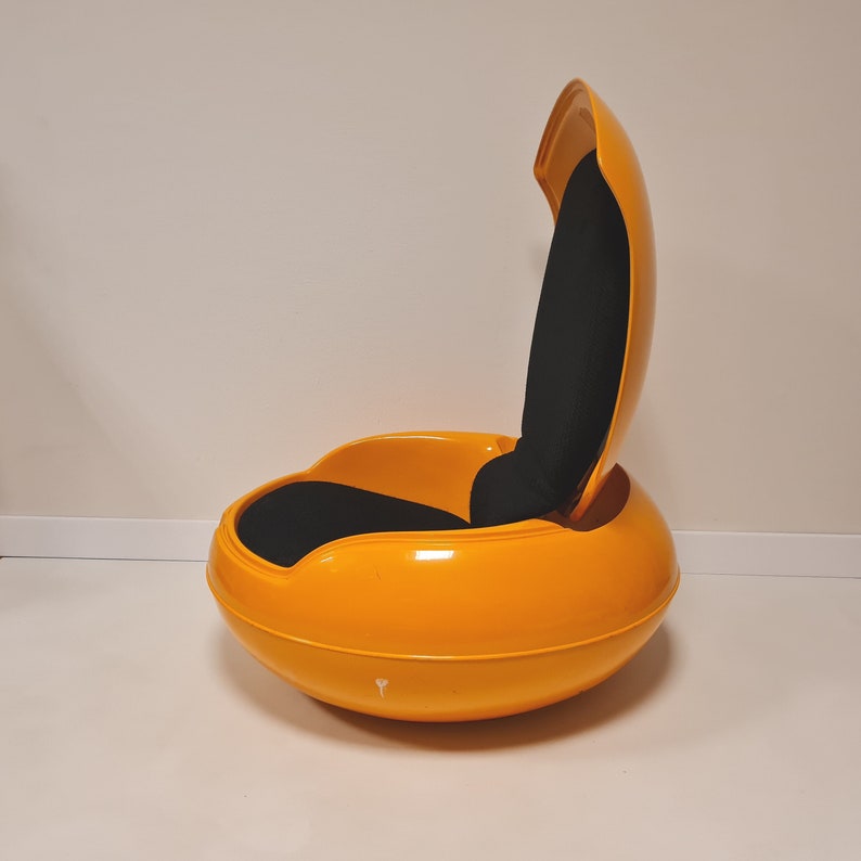 May include: A bright yellow egg-shaped chair with a black fabric cushion and a hinged back. The chair is made of plastic and has a smooth, glossy finish.