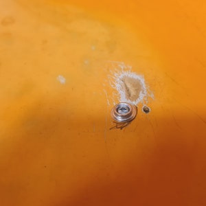 May include: Close-up of a silver screw with a round head, mounted on a bright orange surface. The surface has some scratches and a small white mark.