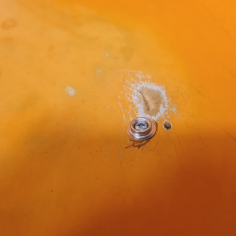 May include: Close-up of a silver screw with a round head, mounted on a bright orange surface. The surface has some scratches and a small white mark.