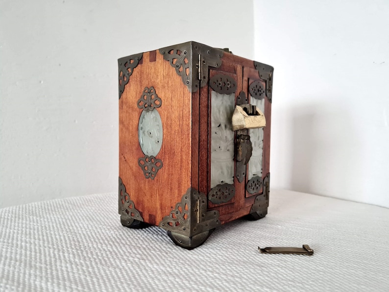 May include: A small wooden box with a hinged lid and a metal lock. The box is decorated with intricate metalwork and has a jade-colored inlay. The box is sitting on a white surface.