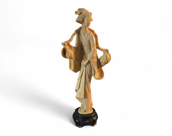 May include: A beige figurine of a woman in traditional Japanese attire, holding two baskets. The figure stands on a black, ornate base. The woman's kimono and hairstyle are detailed, and the overall aesthetic is elegant.