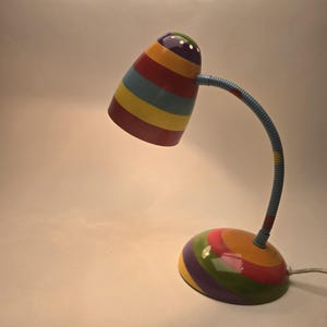 May include: A colorful desk lamp with a rainbow striped shade and base. The lamp has a flexible, blue gooseneck arm and a round base. The shade is cone-shaped with a series of colored stripes. The lamp is plugged into an electrical outlet.