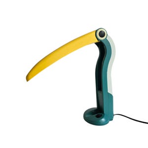 May include: A whimsical desk lamp in the shape of a toucan's beak. The lamp features a curved, yellow light arm, a teal base and body, and a white accent. The lamp is plugged in with a black cord.