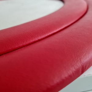 May include: Close-up of a red leather steering wheel cover. The cover has a smooth, textured surface and a double-stitched seam.