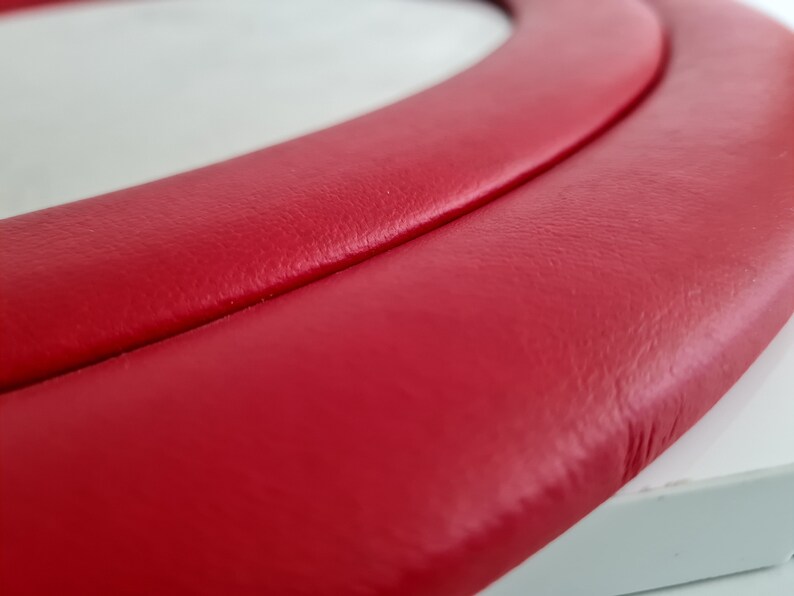 May include: Close-up of a red leather steering wheel cover. The cover has a smooth, textured surface and a double-stitched seam.