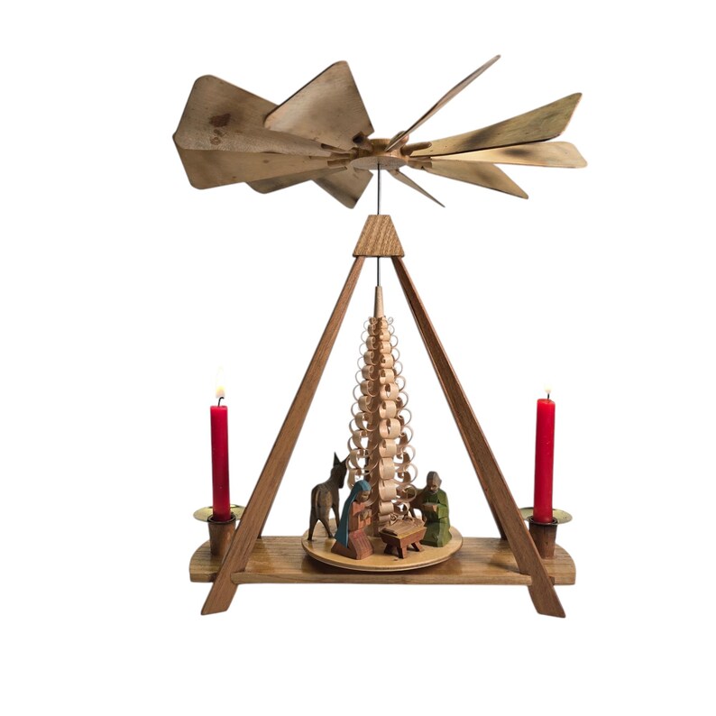 May include: A wooden Christmas pyramid with a rotating, layered tree in the center. The pyramid has a gold star on top and two red candles on the sides. The base of the pyramid features a nativity scene with figures of Mary, Joseph, and a donkey.