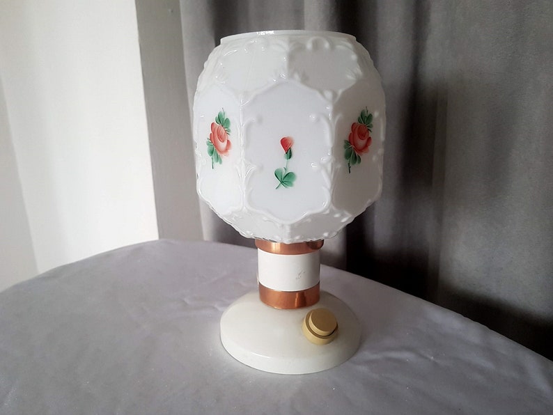 May include: White table lamp with a white glass shade featuring a floral design with red roses and green leaves. The lamp has a copper base and a white switch.
