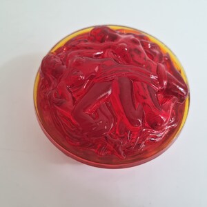 May include: A red glass bowl with a raised design of intertwined figures. The bowl has a yellow rim.