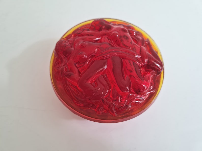 May include: A red glass bowl with a raised design of intertwined figures. The bowl has a yellow rim.
