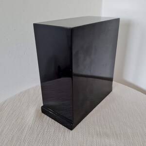 May include: A black wooden box with a smooth, glossy finish. The box has a simple, rectangular shape and a slightly raised base.