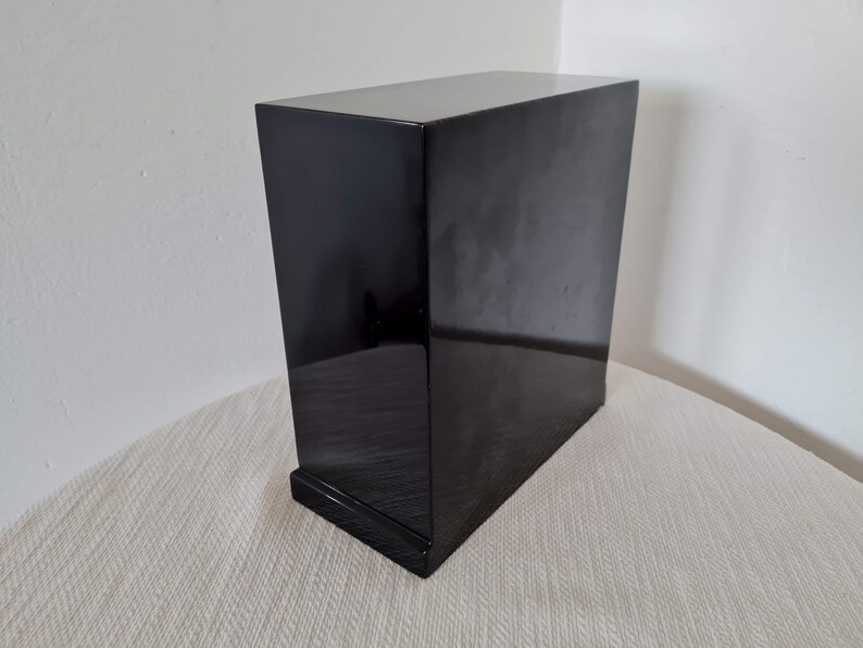 May include: A black wooden box with a smooth, glossy finish. The box has a simple, rectangular shape and a slightly raised base.