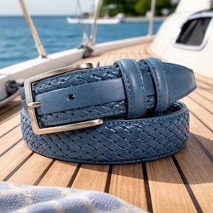 May include: A blue woven leather belt with a silver buckle. The belt is coiled on a wooden surface, with a glimpse of a blue ocean and a boat in the background. The belt's texture is visible, showcasing its intricate design.