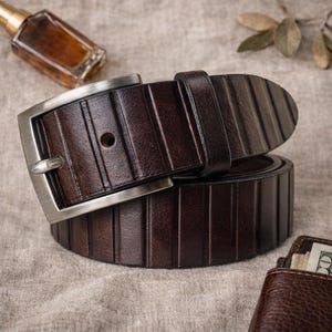 Op de afbeelding: Een donkerbruine leren riem met een zilveren gesp. De riem heeft verticale geribbelde details en is opgerold. De gesp is rechthoekig met een enkele pin. De riem is waarschijnlijk 3,18 cm breed.