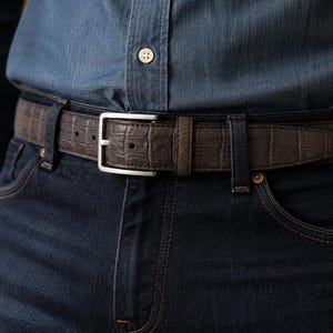 Puede incluir: Un cinturón marrón oscuro con un patrón texturizado de relieve de cocodrilo y una hebilla rectangular de metal plateado. El cinturón se lleva a través de las trabillas de unos vaqueros azul oscuro, bajo una camisa azul con un botón visible.