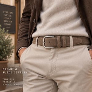 May include: A taupe suede leather belt with a silver-toned metal buckle, worn with beige trousers, a light-coloured jumper, and a dark brown jacket. The image includes the text: ITALIAN STYLE FOR A BIGGER STORY, PREMIUM SUEDE LEATHER, TIMELESS, VERSATILE, MADE TO LAST.