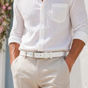 May include: A white linen shirt with a chest pocket, paired with beige linen pants and a white leather belt. The outfit is completed with a silver buckle. The shirt is buttoned up and the sleeves are rolled up.
