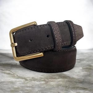 May include: A brown suede belt with a gold-coloured buckle. The belt features a classic design with a rectangular buckle and a loop to secure the belt end. The belt is made of a soft, textured material.