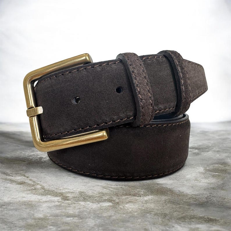 May include: A brown suede belt with a gold-coloured buckle. The belt features a classic design with a rectangular buckle and a loop to secure the belt end. The belt is made of a soft, textured material.
