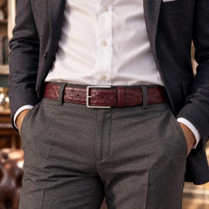 Puede incluir: Un cinturón de color burdeos profundo con un patrón texturizado similar al avestruz y una hebilla de metal plateado, usado sobre pantalones grises con una camisa blanca abotonada y un blazer gris oscuro. El cinturón tiene un cierre de hebilla.