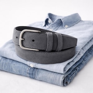 Può includere: Una cintura grigia in pelle scamosciata con fibbia argentata è appoggiata su una pila di camicie di jeans blu piegate. La cintura ha un design classico con fibbia ad ardiglione e due passanti. Le camicie sono di diverse tonalità di blu.