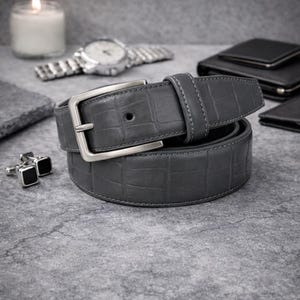 May include: A grey leather belt with a silver buckle. The belt has a textured, crocodile-like pattern. Other accessories include a watch, wallet, and cufflinks, all in shades of grey and black. The belt is a stylish accessory.