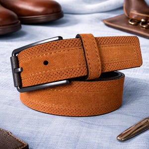May include: A tan suede belt with a black buckle. The belt features a perforated pattern along the edges and a single hole for the buckle. The buckle is rectangular and has a matte finish. The belt is likely made of genuine leather.
