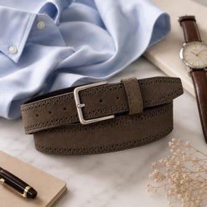 Pode incluir: Um cinto de camurça em tom taupe com perfurações decorativas estilo brogue ao longo das bordas e uma fivela retangular de metal prateado. O cinto está enrolado próximo a uma camisa azul clara e um relógio de pulso com pulseira de couro marrom.