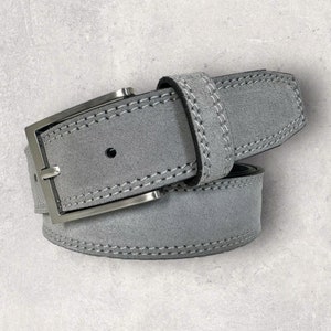 May include: A grey suede belt with a silver rectangular buckle. The belt has white stitching along the edges and around the buckle. The belt is a neutral colour, suitable for various outfits.